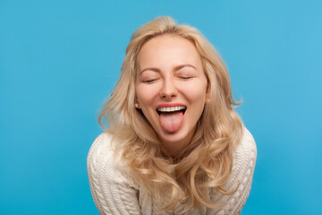 Closeup naughty playful woman with blond curly hair showing tongue at camera with closed eyes, having fun, joke. Indoor studio shot isolated on blue background