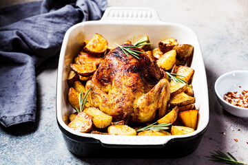 Baked whole chicken with potatoes and rosemary in black dish, dark background.