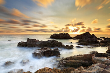 Obraz premium Long exposure of sea wave impact the stone with clouds moving on beach in the sunset time, Eastern of Thailand