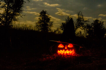 Carved halloween pumpkin jack-o-lantern wearing witch hat with burning candles glows in a darkness. Spooky landscape
