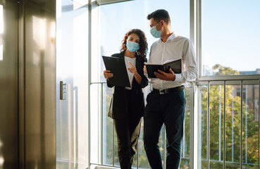 Business colleagues in medical face mask discussing together work issues standing near elevator at office. Meeting near elevator. Teamwork during pandemic in quarantine city. Covid-19.