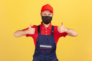 Positive crafts man in red and blue uniform and protective facial mask showing thumbs-up, satisfied with quality service during quarantine. Indoor studio shot isolated on yellow background