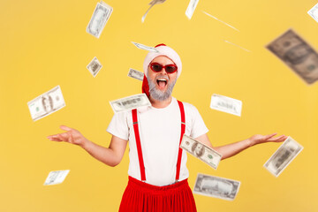 Extremely smiling positive adult man in santa claus hat and trendy sunglasses enjoying standing under dollar banknotes rain, lottery victory, income. Indoor studio shot isolated on yellow background