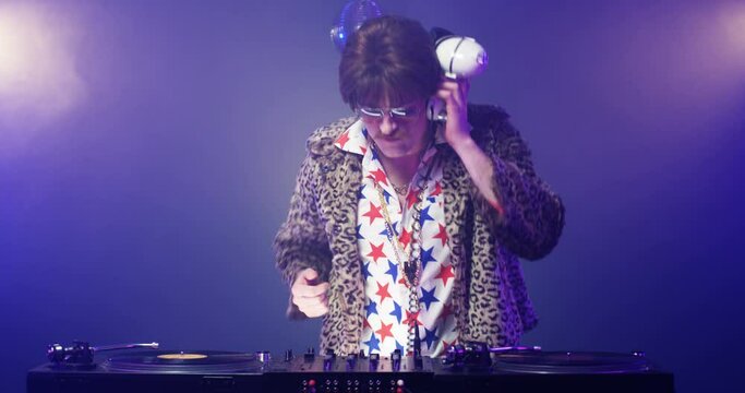 Stylish Caucasian vintage man touching vinyl and dancing while mixing sounds on equipment at party. Retro style male DJ in leopard jacket with headphones playing music at nightclub. Disco concept