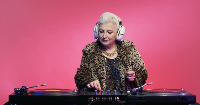 Retro style Caucasian old woman in headphones and leopard coat playing music as dj on pink background. Vintage senior female dancing while mixing on audio equipment. Stylish clubbing grandma.