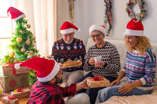 Group Of Four Mature And Young People Having Fun At Home The Christmas Day Wrapping The Presents And Gifts Together - Man Giving At His Woman A Gift