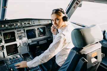 Woman pilot sitting in aircraft cockpit, wearing headset, looking at camera. - obrazy, fototapety, plakaty