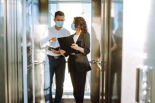 Business partners in protective sterile mask in the elevator. Colleagues talking while standing in elevator at  modern office. Teamwork during pandemic in quarantine city. Covid-19.