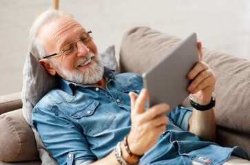 Cheerful aged man using tablet on couch.