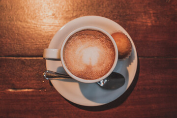 Taza de café con leche en un plato blanco sobre una mesa de madera con una cuchara de plata y un miini cupcake