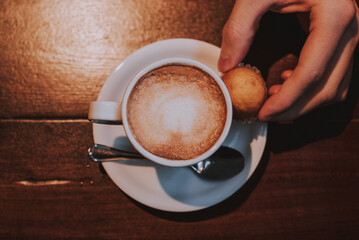 Taza de café con leche en un plato blanco sobre una mesa de madera con una cuchara de plata  y una mano sujetando un cupcake