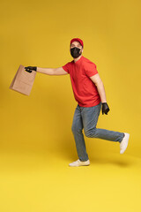 Fototapeta na wymiar Man in red cap, t-shirt in protective mask and gloves giving fast food order isolated on yellow background. Male employee courier hold empty paper packet with food