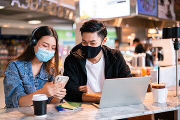 Asian couple take off mask protection uses their laptop computer and smartphone to relax during a flight in lounge at international airport.Young man and woman laugh smile together in coffee shop cafe