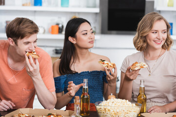 joyful multicultural friends eating pizza near bowl of popcorn