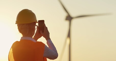 Back view on the caucasian woman in the helmet taking photos of the big windmill tourbine producing...