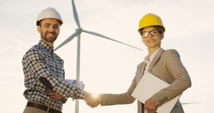 Caucasian Man Engineer And Woman Environmental Consultant In Helmets Shaking Hands And Smiling To The Camera On The Windmill Turbine Background. Portrait.