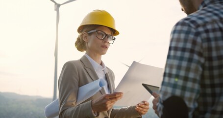 Woman in a helmet, environmental observer with documents in hands standing together with a male worker of the windmill power station and giving him orders and advice while he noting on the tablet.