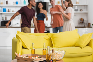 selective focus of beer and pizza on table near yellow sofa and multicultural friends dancing on background