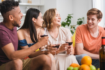 excited multicultural friends talking while holding glasses of red wine in kitchen