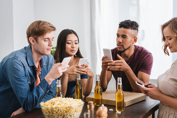selective focus of multicultural friends chatting on smartphones near beer and popcorn during party