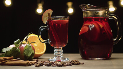 Close up hot red mulled wine in a glass with slice of apple and cinnamon stick. Jug with milled wine. Blurry lights on black background