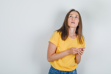 Young woman in t-shirt, shorts looking up while holding smartphone and looking pensive , front view.