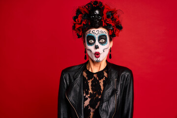 Scary young creepy lady calavera. wears artistic make-up for the feast of all the dead. Has frightened expression, wears black leather jacket and lace gloves, dressed as skeleton isolated in red.