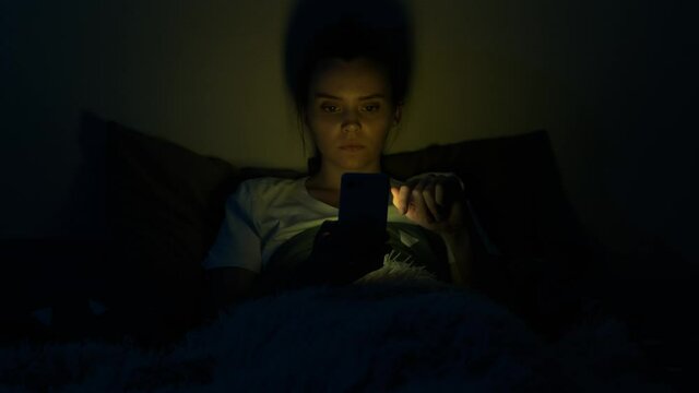 Girl using mobile phone lying in bed at night. Young woman browsing online and scrolling social media in smart phone