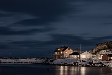Wooden cottage on the shore of the sea at night in winter. View of harbour with wooden houses in winter season surrounded by snowy mountains © Alena