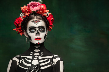 Scary. Young girl like Santa Muerte Saint death or Sugar skull with bright make-up. Portrait isolated on dark green studio background with copyspace. Celebrating Halloween or Day of the dead.