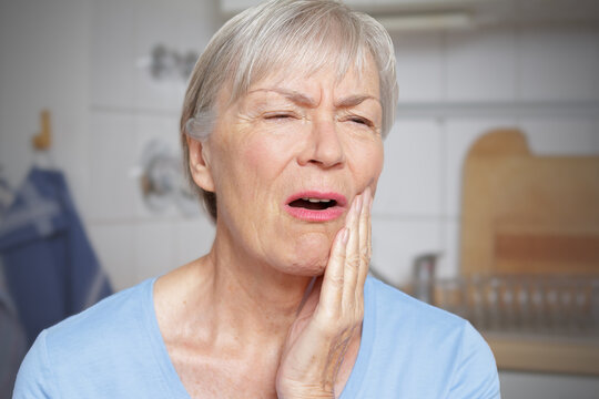 Acute toothache: elderly woman in her kitchen with a hand at her painful cheek.