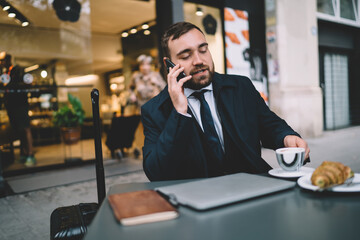 Formally dressed male owner sitting at cafe table with closed laptop device and making international smartphone conversation during breakfast time, Caucasian boss using mobile for roaming phoning
