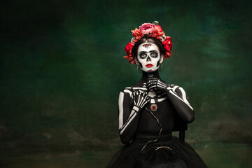 Poison. Young girl like Santa Muerte Saint death or Sugar skull with bright make-up. Portrait isolated on dark green studio background with copyspace. Celebrating Halloween or Day of the dead.