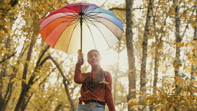 Cheerful teenage girl opens an umbrella above herself in an autumn park, leaves are falling down, slow motion
