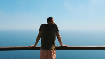 Young man on vacation comes to the railing, clouds on it and enjoys beautiful scenic view of the sea or ocean with ships in the distance. The guy looks at the ocean, follow up shot
