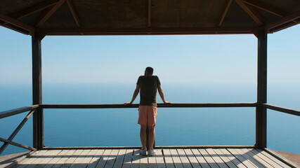 Young man on vacation comes to the railing, clouds on it and enjoys beautiful scenic view of the sea or ocean with ships in the distance. The guy looks at the ocean, follow up shot