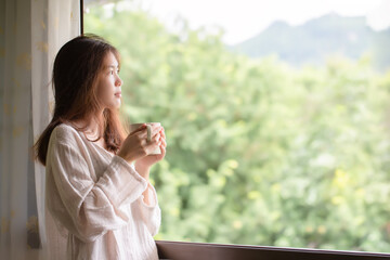 Asian woman drinking coffee in the morning sunlight after waking up in her bedroom.