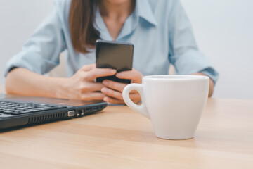 Fototapeta na wymiar White hot coffee mug on the desk office and female employee using mobile phone on background.