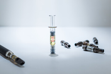 A THC distillate syringe standing upright, in the middle of used vape cartridges, and a vape pen on...