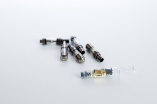 A pile of used disposable vape cartridges behind a full distillate syringe, ready for a reusable cartridge.  Shows the waste that can be saved by using refillable cartridges.
