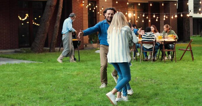 Caucasian happy cheerful father and mother running and playing with kids on green grass in yard. Family barbeque on background. Joyful children with dad and mom having fun and laughing. Outside.
