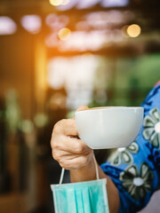 Hot Cappuccino coffee with protective mask to prevent Corona Virus (Covid-19) hanging on woman arm while take off mask to drink coffee at coffee shop. New normal lifestyle concept. Selective focus