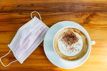 Hot Cappuccino coffee with protective mask on a wooden table for people being quarantined in the...