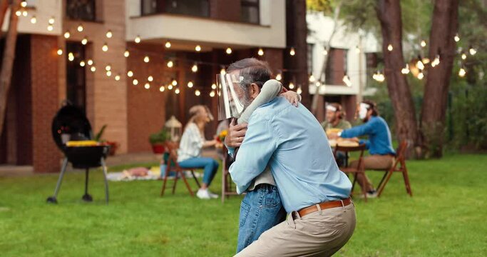 Caucasian happy grandfather and small cute granddaughter in medical face shields hugging in yard. Family at table with barbeque dinner on background. Senior grandpa embracing little girl in masks.