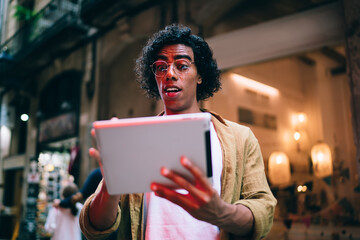 Handsome curly hipster guy in eyewear reading news and stressed with updates on digital tablet, handsome young millennial male 20s watching sholing video on touchpad standing outdoors near neon lights