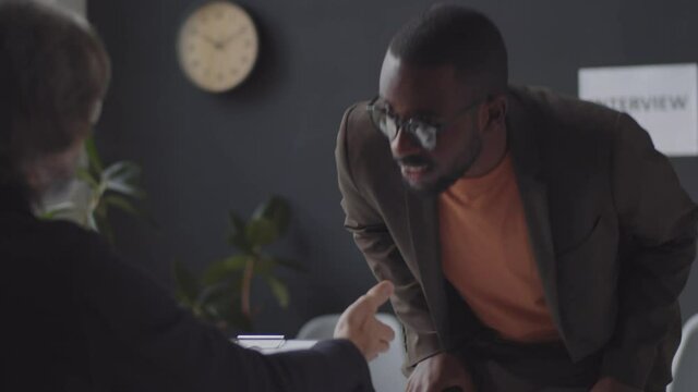 Young African American man greeting HR manager with handshake, giving CV to him and answering questions while having job interview in office