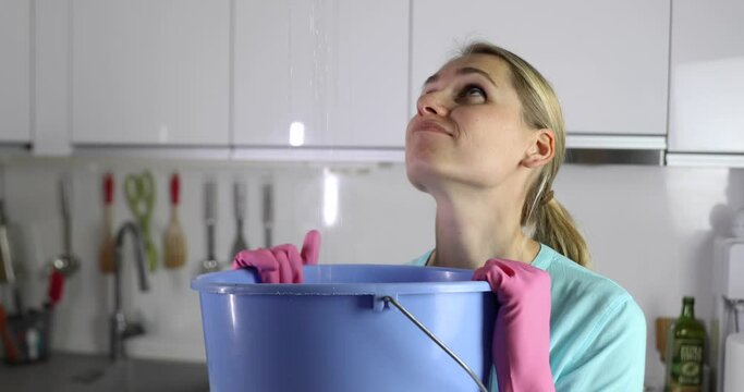 home flooded by upstairs or roof damage - woman holding bucket while water leaking from ceiling in kitchen