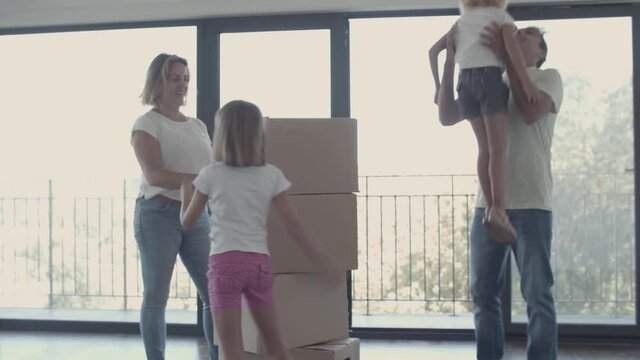 Parents and two girls celebrating moving into new apartment, carrying cartoon boxes, making stack and hugging. Dolly shot. Moving into new house concept