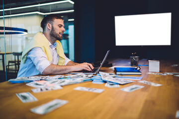 Caucasian male investor in smart casual wear sitting at coworking table with modern technology for making money during sports betting, businessman checking income earnings in room with blank TV screen