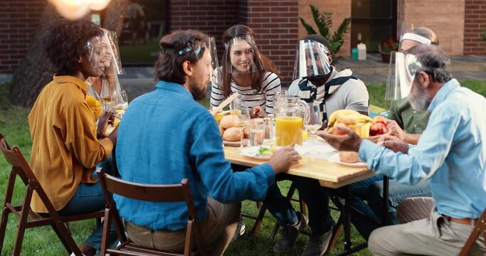 Mixed-races people in face shields sitting at dinner table in back yard and talking. Multi ethnic family having picnic meal in court and chatting friendly. Communication of generations during pandemic